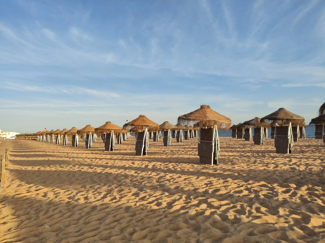 Rows of straw beach umbrellas and wooden loungers on a shallow sandbar with people wading in clear water.