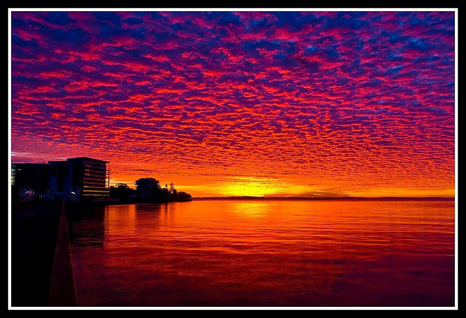 A wooden jetty at sunset stretching over calm Red Sea water, with soft orange light reflecting on the surface and silhouettes of distant mountains.